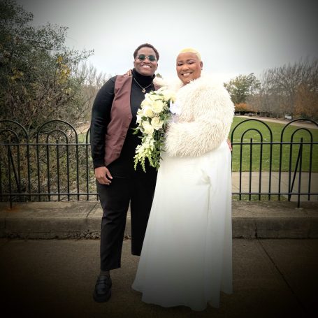 Two women standing together outdoors, one in a wedding dress holding flowers, smiling. Boundless Love.