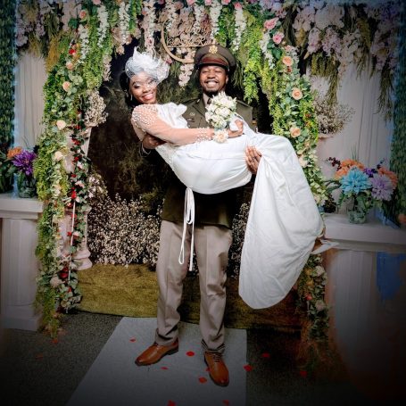 A happy couple poses in wedding attire, surrounded by flowers and greenery. Convenient Location.